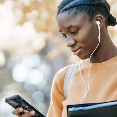 Positive young African American female student with earphones and folder using mobile phone in park